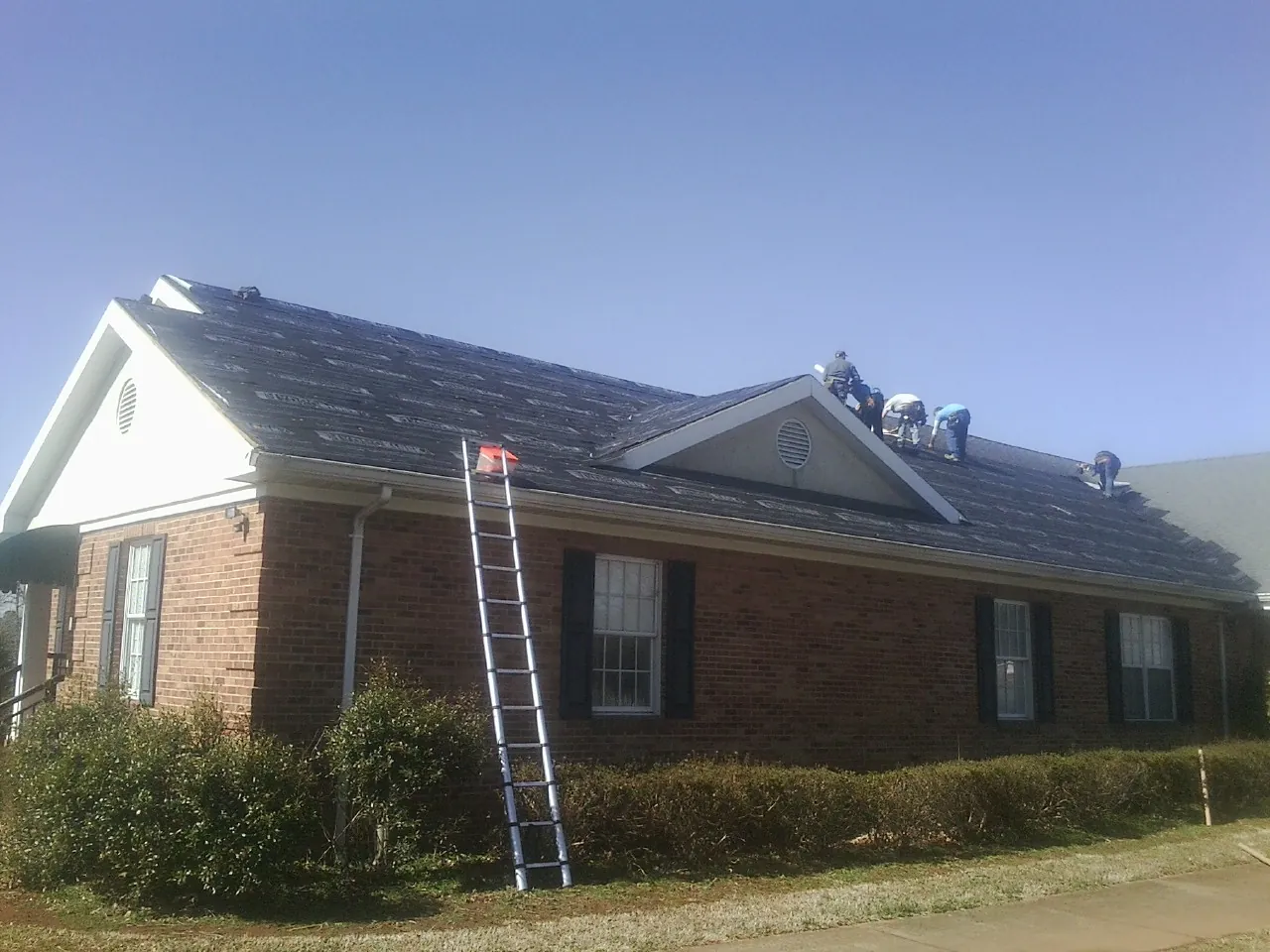 Crew tearing shingles off a brick ranch roof with a ladder set — Emergency Roof Repair work by our crew in Social Circle