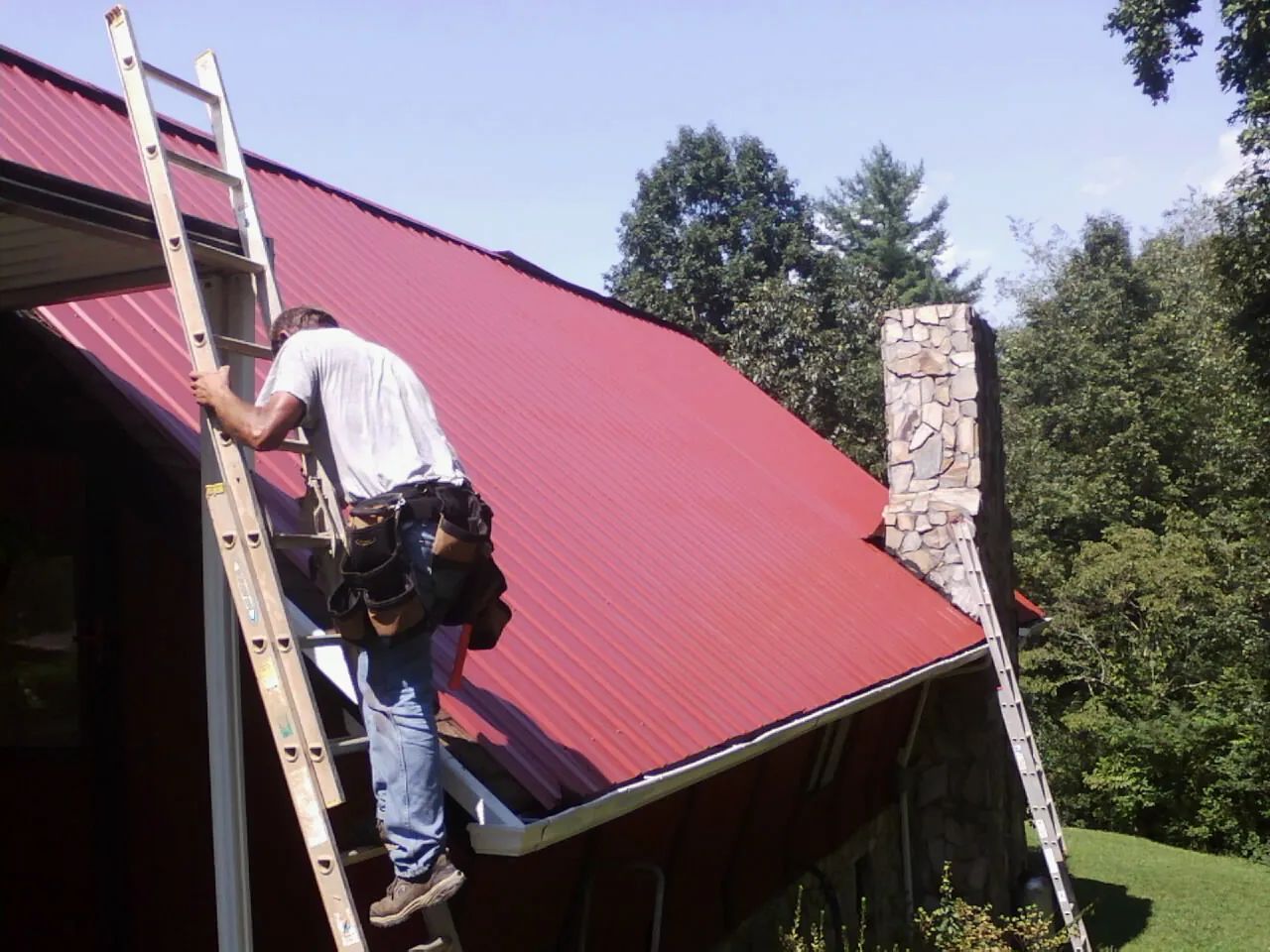 Roofer on a ladder beside a red metal roof and stone chimney — Soffit & Fascia Repair by Roofing Brothers in Social Circle
